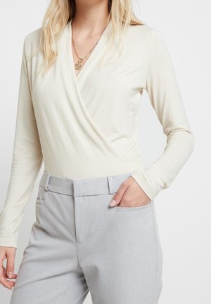 Kobieta ubrana w kremową, kopertową bluzkę z długim rękawem oraz jasnoszare, wysokie spodnie z jedną ręką w kieszeni.