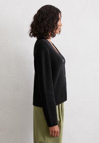 Schwarze gestrickte Strickjacke mit V-Ausschnitt, zwei vorderen Knöpfen und langen Ärmeln, kombiniert mit grünen Hosen. Strukturierter und kurz geschnittener Stil.