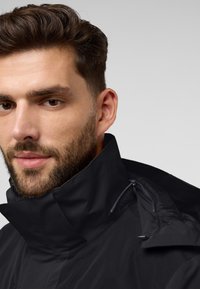 Veste noire imperméable avec un col haut, des poches zippées et une texture lisse. Présente un design minimaliste avec des accents gris.