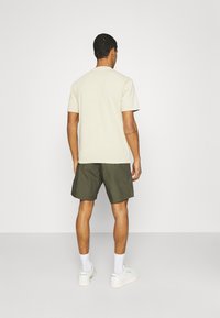 Homme debout de dos, portant un t-shirt beige uni, un short vert foncé, des chaussettes blanches et des sneakers Reebok blanches.