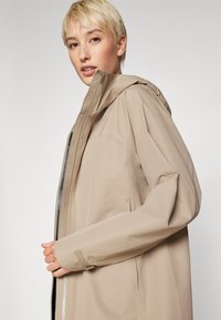 Beige, vedenpitävä takki huppulla, edessä vetoketjulla, säädettävillä hihansuilla ja rennolla leikkauksella. Sujuva pinta, minimaalinen muotoilutyyli.