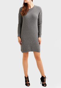 Graues Strickkleid mit Rautenmuster, Rundhalsausschnitt und langen Ärmeln. Kombiniert mit schwarzen Schnürheels. Glatte Textur.