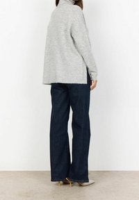 Pull gris en maille avec un col montant, coupe ample et fentes latérales, associé à un jean larges bleu foncé et des talons argentés pointus.