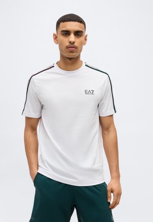 Fiatal férfi, fehér EA7 Emporio Armani pólóban, fekete és zöld csíkos vállrészlettel és sötétzöld sportos rövidnadrágban.