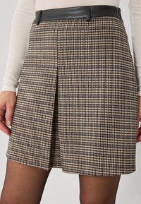 Egy bézs, barna és fekete kockás szoknya sima fekete derékrésszel és elülső redővel, oldalt lévő kéz mellett bemutatva.