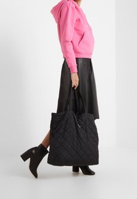 Bolso tote negro acolchado con cierre de cordón, superficie texturizada y correas anchas, sostenido por una persona con sudadera rosa con capucha y falda negra.
