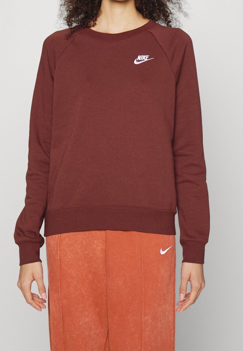 Personne portant un sweat-shirt Nike bordeaux et un pantalon Nike orange, tous deux avec des logos swoosh blancs visibles, debout devant un fond uni.