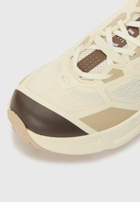 Helle beige Laufenschuhe mit einem strukturierten Mesh-Obermaterial, einer braunen Gummispitze und Schnürsenkeln über einer gepolsterten Zunge mit einem gemusterten Akzent.