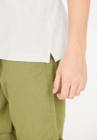 Vit, kortärmad skjorta med slits på sidan; matchad med olivgröna, texturerade byxor. En hand vilar på byxorna.