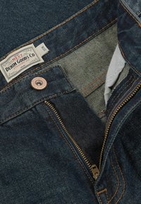 Vaqueros de mezclilla azul oscuro con costuras naranjas en contraste, cierre de cremallera y una etiqueta tejida que muestra "DENTAL GOODS CO, AJUSTE RECTO."