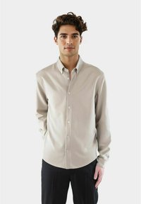 Ung mann med krøllete hår iført en beige skjorte med knapper og svarte bukser, stående mot en ensfarget lys bakgrunn.