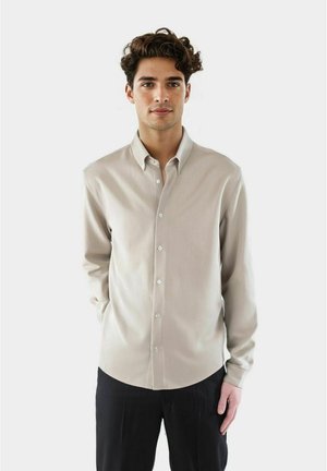 Ung mann med krøllete hår iført en beige skjorte med knapper og svarte bukser, stående mot en ensfarget lys bakgrunn.