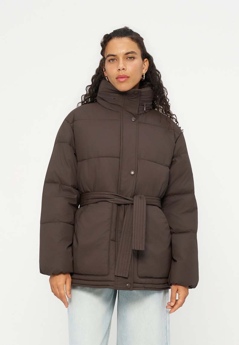 Femme aux cheveux bouclés portant une doudoune marron foncé ceinturée et un jean bleu clair, debout devant un fond clair uni.