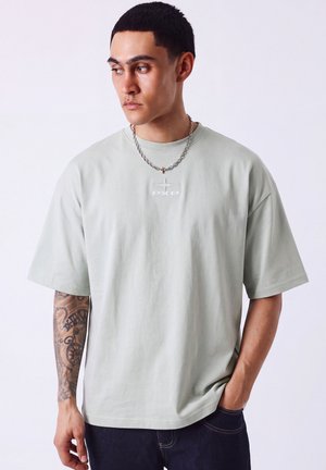 Giovane uomo con capelli corti e neri che indossa una t-shirt oversize grigio chiaro con il logo PXP, jeans scuri, collana a catena e tatuaggio sul braccio, guarda a sinistra.
