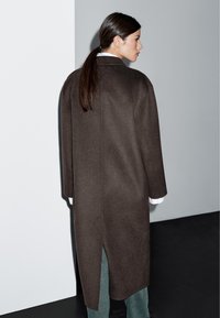 Długi brązowy wełniany płaszcz o wyraźnie zarysowanym kształcie, z bocznymi rozcięciami i pełnymi rękawami, noszony na białej koszuli z kołnierzykiem i zielonych spodniach.