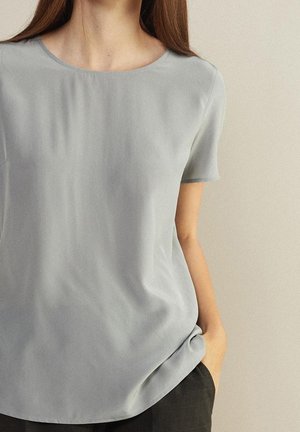 Kvinde iført en ensfarvet lysegrå kortærmet bluse og sorte bukser, stående med den ene hånd i lommen mod en lys væg.