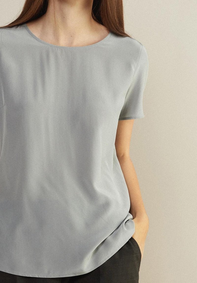 Femme portant un chemisier uni gris clair à manches courtes et un pantalon noir, debout avec une main dans sa poche, appuyée contre un mur clair.