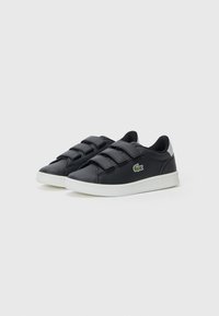 Černé kožené tenisky se třemi suchými zipy, bílé gumové podrážky a malým zeleným logem Lacoste na boku. Hladký povrch, neformální design.