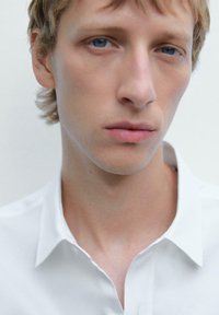 Weißes Hemd mit glatter Textur, getragen von einer Person mit kurzen, hellen Haaren und einem neutralen Hintergrund, der auf die Gesichtszüge fokussiert.