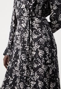 Crna haljina od tkanine s bijelim cvjetnim uzorcima, dugim rukavima, vezivanjem u struku i opuštenim krojem s suptilnim nabranim detaljima.