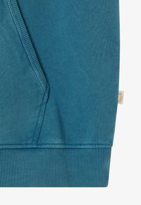 Skarpetka w kolorze niebiesko-skalnym z widocznymi szwami, prążkowanym dołem i małą beżową metką po prawej stronie z napisem "BLK WVS".