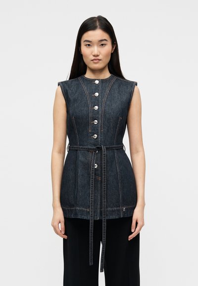 Femme aux longs cheveux noirs portant un gilet sans manches en denim foncé boutonné avec une ceinture à la taille, sur un pantalon noir, regardant vers l'avant sur un fond uni.