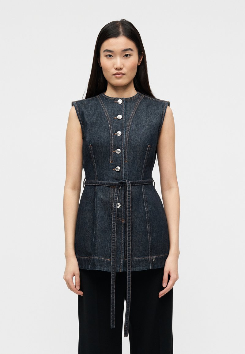 Γυναίκα με μακριά σκούρα μαλλιά πο υ φορά αμάνικο σκούρο τζιν γιλέκο με κουμπιά και ζώνη στη μέση πάνω από μαύρο παντελόνι, κοιτάζοντας προς τα εμπρός με φόντο απλό.