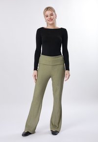 Νεαρή γυναίκα στέκεται, φοράει μαύρο μακρυμάνικο τοπ, ψηλόμεσα ελαστικά παντελόνια σε ελιά πράσινο χρώμα και μαύρα επίπεδα παπούτσια, χαμογελώντας στην κάμερα.