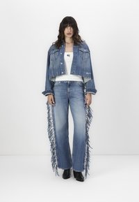 Denimjack met een cropped ontwerp, franjes op de randen en twee voorzakken, gecombineerd met een wijde jeans met franjes aan de zijkanten. Zwarte laarzen.