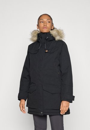 Vrouw in een zwarte winterparka met een met bont gevoerde capuchon, staande tegen een effen lichte achtergrond, met een lichte blik opzij.