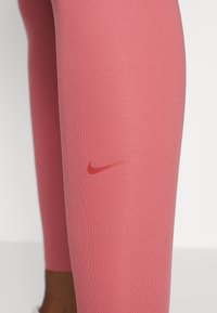 Legginsy Nike w intensywnym różowym kolorze, charakteryzujące się gładką teksturą oraz małym czerwonym logo swoosh z boku. Zbliżenie na detal nogawki.