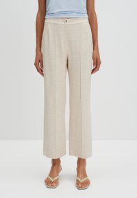 Beige wijde broek met een gestructureerde stof, voorzien van een sluiting met een knop aan de voorkant en twee verticale plooien. Gecombineerd met simpele beige sandalen.