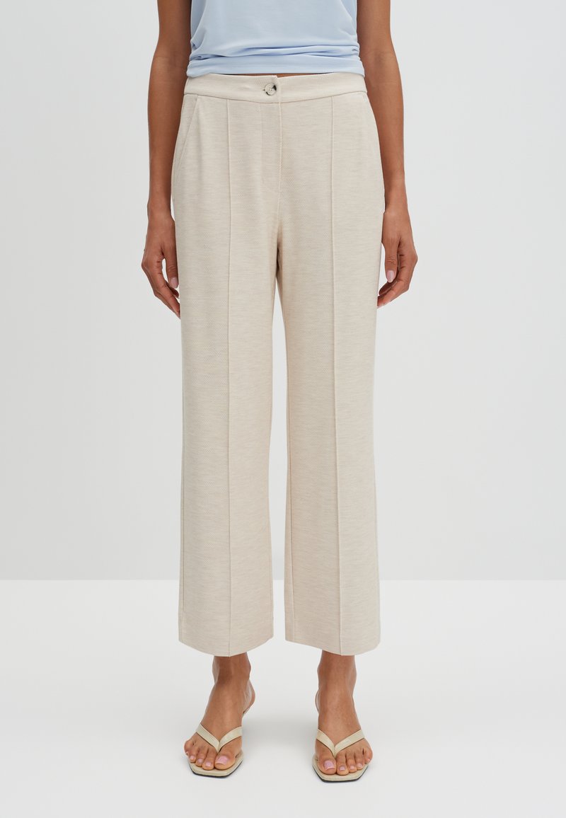 Beige wijde broek met een gestructureerde stof, voorzien van een sluiting met een knop aan de voorkant en twee verticale plooien. Gecombineerd met simpele beige sandalen.