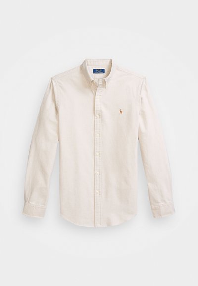 Kremkrāsas pogātā krekls, kas izgatavots no kokvilnas. Ievērojams ar klasisku apkakli, garām piedurknēm un izšūtu logotipu uz krūtīm.