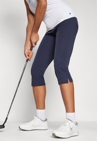 Pantalones capri de golf en color navy con aberturas laterales, usados con zapatillas deportivas blancas y calcetines cortos, combinados con una blusa blanca. Presenta un diseño ajustado.
