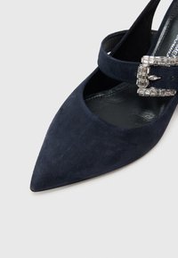 Γόβα σε σκούρο μπλε σουέντ με μυτερή μύτη και διακοσμητικό μεταλλικό λουράκι με αγκράφα. Η λεία υφή και η κομψή σιλουέτα αναδεικνύουν το σχέδιο.