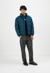 Joven de pie llevando una chaqueta acolchada color teal, pantalones grises con estampado, zapatos negros, con las manos en los bolsillos, contra un fondo blanco liso.