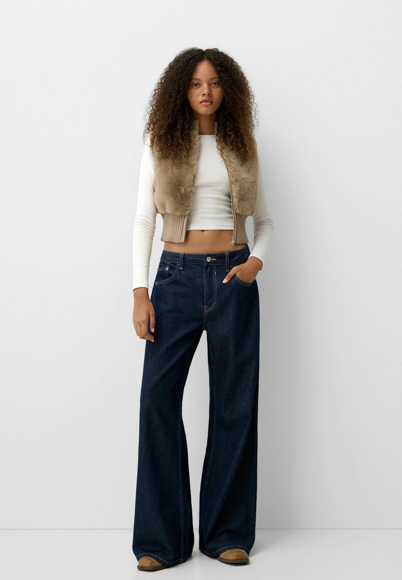 Frau mit lockigem Haar, die eine beige Pelzweste über einem weißen Crop-Top, dunklen, weiten Jeans und braunen Schuhen trägt und vor einem weißen Hintergrund steht.