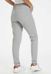 Persoon in lichtgrijze joggingbroek met elastische tailleband en witte sneakers met bruine hielaccenten, staande met handen in de zakken.