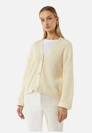 Cremfarbener Strickcardigan mit V-Ausschnitt, oversized Passform, vier Knöpfen auf der Vorderseite und weiten Ärmeln. Getragen über einem weißen Hemd.