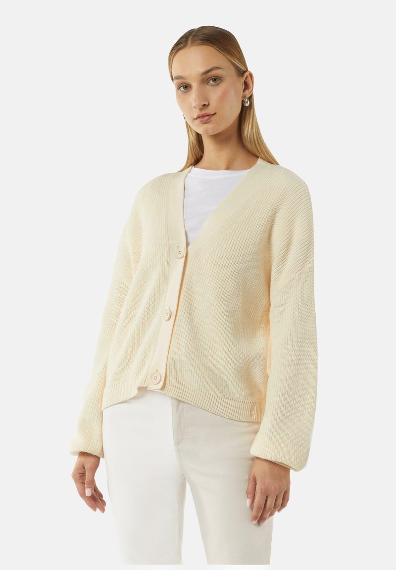Cremfarbener Strickcardigan mit V-Ausschnitt, oversized Passform, vier Knöpfen auf der Vorderseite und weiten Ärmeln. Getragen über einem weißen Hemd.
