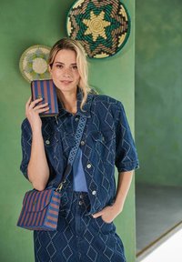 Denim jack met een diamantpatroon, gecombineerd met een blauw geribbeld topje en een gestreepte blauw-rode schoudertas. Groene achtergrond.