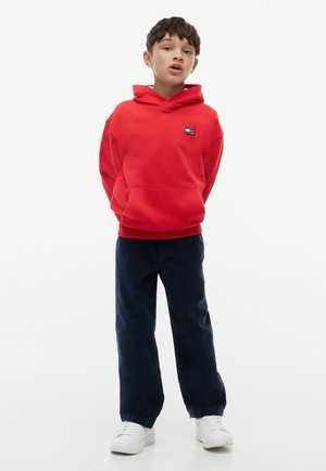 Dreng iført en rød hættetrøje, mørkeblå bukser og hvide sneakers, der står med hænderne bag ryggen mod en hvid baggrund.