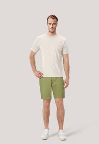 T-shirt in cotone beige abbinato a pantaloni corti verdi e sneakers bianche. La maglietta ha maniche corte, mentre i pantaloni corti sono lunghi fino al ginocchio con una texture liscia.