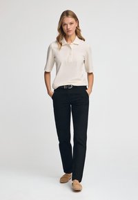 Beige polo met korte mouwen en knoopsluiting, gecombineerd met zwarte broeken en tan instappers. De outfit heeft een gladde textuur en een verfijnde pasvorm.