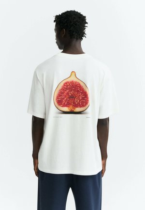 Persona con pelle scura e capelli ricci vista di spalle che indossa una camicia bianca con una grande immagine di un fico sulla schiena.