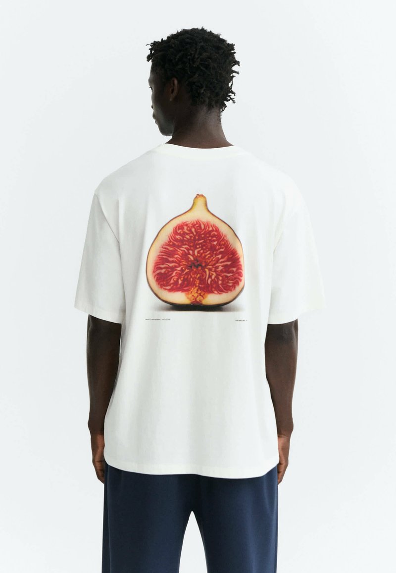 Personne à la peau foncée et aux cheveux bouclés, vue de dos, portant une chemise blanche avec un grand motif de fruit de figue dans le dos.