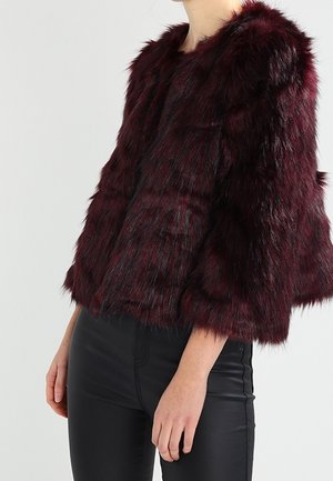 Donna che indossa una giacca corta in pelliccia ecologica bordeaux scuro con maniche a tre quarti e pantaloni neri a vita alta su sfondo semplice.