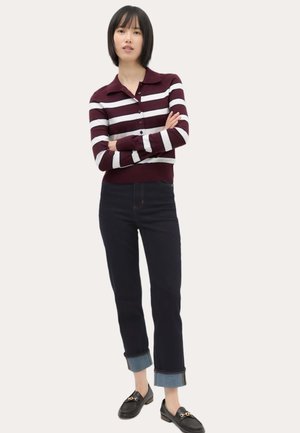 Ung kvinde med kort sort hår iført en vinrød og hvidstribet knap-sweater, mørke opbukkelige jeans og sorte loafers, stående med armene krydset.