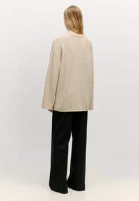 Beige Strukturpullover in Übergröße mit langen Ärmeln und geradem Saum, kombiniert mit schwarzen, weit geschnittenen Hosen. Schlichter Hintergrund.